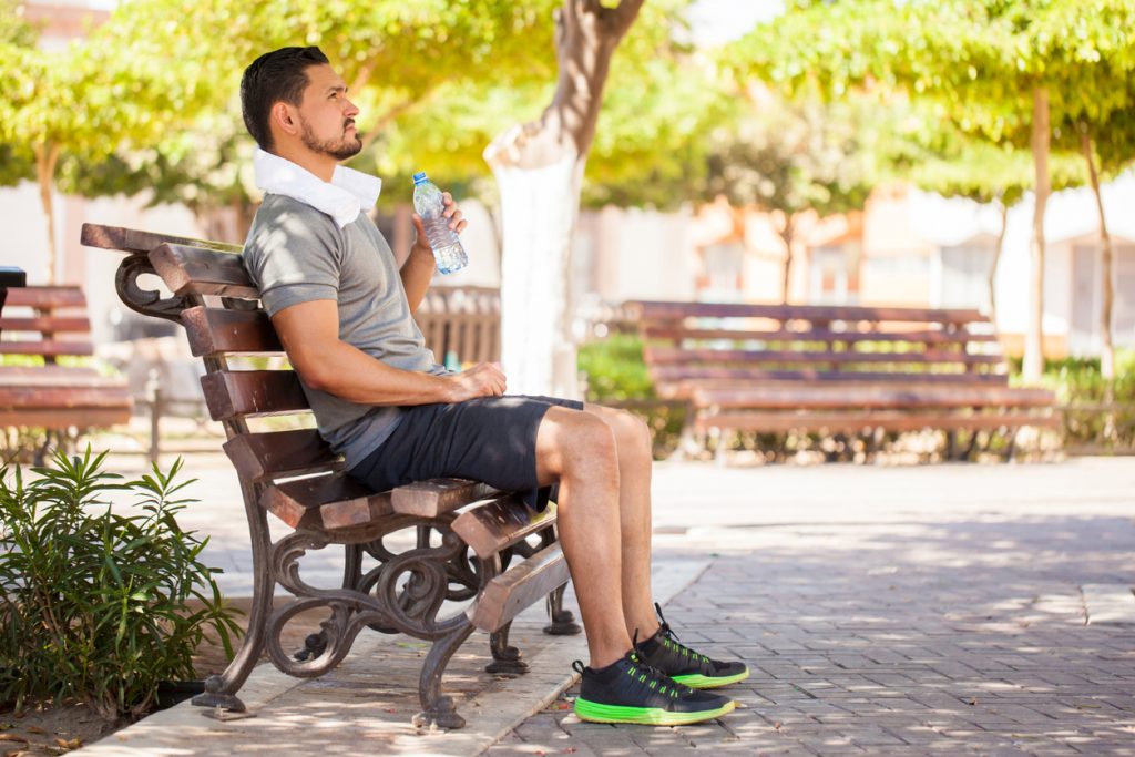 Full length profile view of a Hispanic man with a beard drinking some water from a bottle and resting from working out at a park to stay hydrated.
