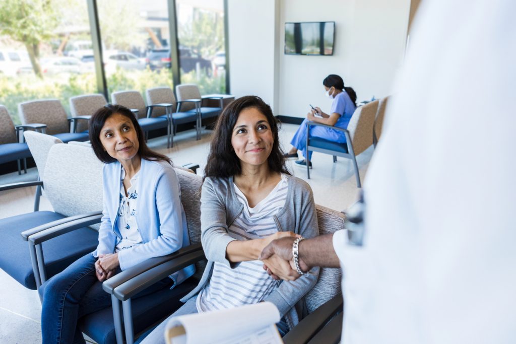 Sitting in the medical waiting room lobby, the mature adult mother watches as her young adult daughter shakes hands with a primary care doctor.