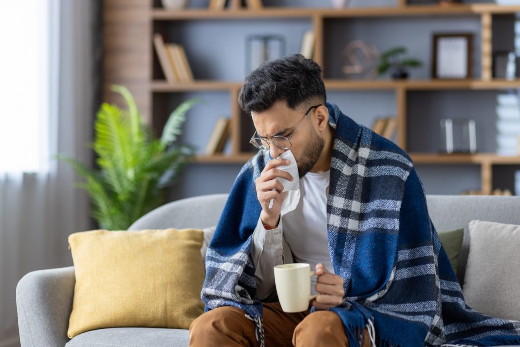 Young man sitting on sofa covered with blanket, holding hot drink and tissue. He appears sick, blowing nose, wrapped in warm blanket, trying to feel better at home during illness or cold season.